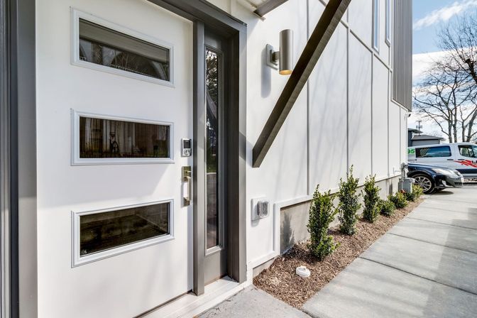 Modern white front door with three horizontal glass panels and smart lock, slim glass sidelight, wall sconce, neat shrub border along a concrete sidewalk and parked cars in the background.