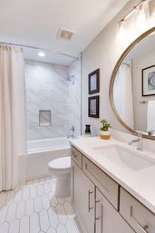 Bright modern bathroom with marble-tiled bathtub and shower, curved shower rod and white curtain, hexagon white floor tiles, gray vanity with quartz countertop, round gold-framed mirror, small potted plant and framed wall art.