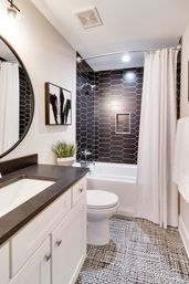 Contemporary bathroom with black hex-tile shower wall, white bathtub and curtain, white vanity with dark countertop, round mirror, patterned floor tile and potted plant.