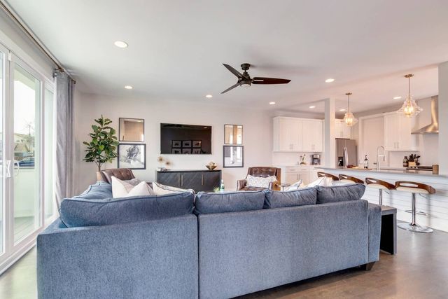 Bright modern open-concept living room and kitchen in a contemporary home — large blue-gray sectional facing a wall-mounted TV and dark media console, leather accent chairs, ceiling fan, sliding glass doors, and a white kitchen island with wooden bar stools and glass pendant lights.