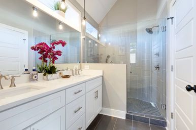 Bright modern master bathroom with white double vanity and quartz countertop, pink orchid centerpiece, large mirror and pendant lights, and a glass-enclosed walk-in shower with gray tile and dark floor tiles