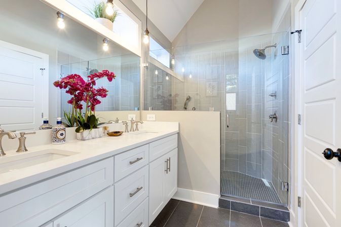 Bright modern master bathroom with white double vanity and quartz countertop, pink orchid centerpiece, large mirror and pendant lights, and a glass-enclosed walk-in shower with gray tile and dark floor tiles