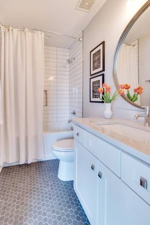 Bright modern bathroom with white vanity and marble countertop, round mirror, vase of orange tulips, subway-tiled shower with white curtain, and gray hexagon tile floor