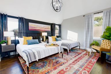 Bright, modern guest bedroom with two twin beds, patterned area rug, vaulted ceiling, large windows, plants and colorful throw pillows.