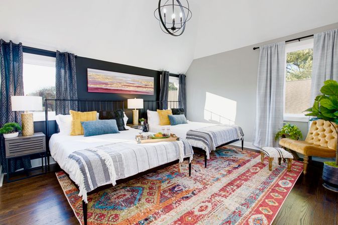 Bright, modern guest bedroom with two twin beds, patterned area rug, vaulted ceiling, large windows, plants and colorful throw pillows.
