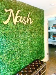 Modern entryway with vibrant faux boxwood accent wall and light wood script sign, tufted leather bench, and open doorway to a bunk-bed bedroom.