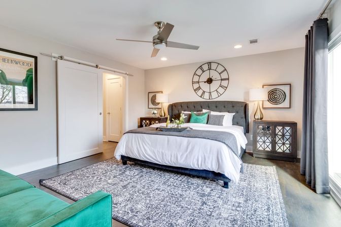 Bright modern master bedroom with king-size bed and tufted gray headboard, oversized wall clock, mirrored nightstands with lamps, sliding barn door, ceiling fan, gray patterned area rug and green accent pillows.