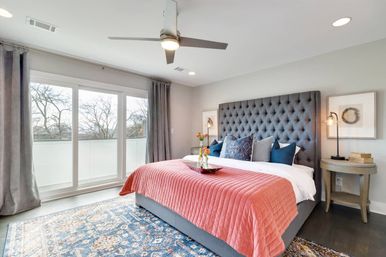 Sunlit modern master bedroom with tufted gray headboard, coral quilted bedspread, navy accent pillows, patterned blue area rug and floor-to-ceiling sliding glass doors opening to a balcony view