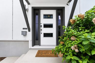Modern suburban front entrance with a white door featuring three horizontal windows and dark trim, concrete steps with a welcome mat, and lush hydrangea bushes with dried blossoms to the right.