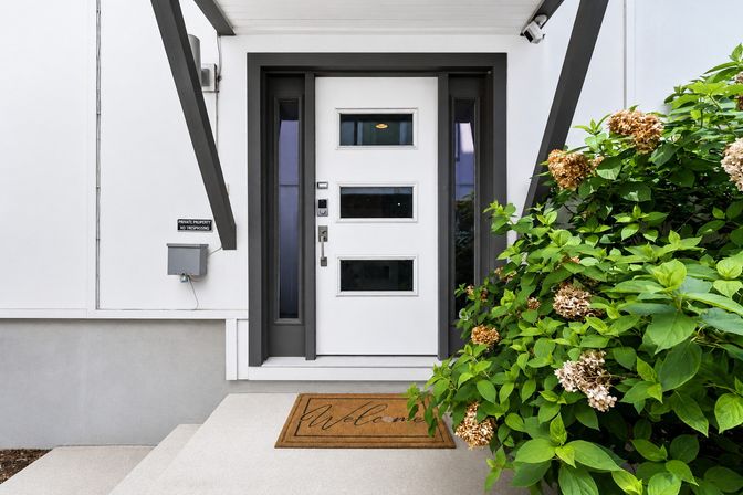 Modern suburban front entrance with a white door featuring three horizontal windows and dark trim, concrete steps with a welcome mat, and lush hydrangea bushes with dried blossoms to the right.