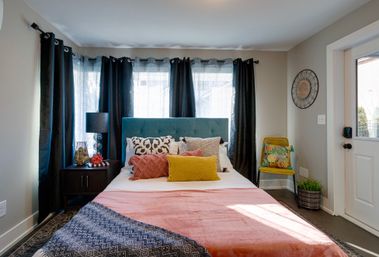 Sunlit modern bedroom with teal upholstered headboard, layered decorative pillows (yellow, coral, patterned), coral throw blanket, dark curtains, nightstand with lamp and decor, yellow accent chair with tropical cushion, potted plant and exterior door.