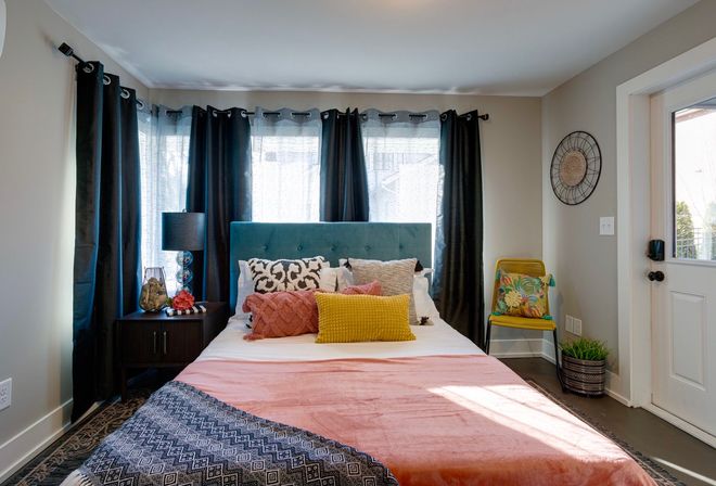 Sunlit modern bedroom with teal upholstered headboard, layered decorative pillows (yellow, coral, patterned), coral throw blanket, dark curtains, nightstand with lamp and decor, yellow accent chair with tropical cushion, potted plant and exterior door.
