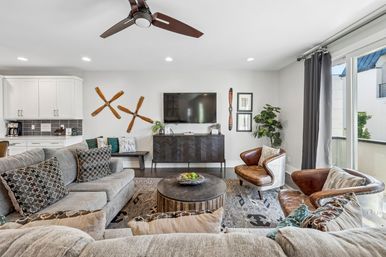 Cozy contemporary living room with a gray sectional, patterned pillows, two leather accent chairs, round wooden coffee table with fruit bowl, wall-mounted TV, wooden propeller wall decor, indoor plant and sliding glass doors to a balcony.