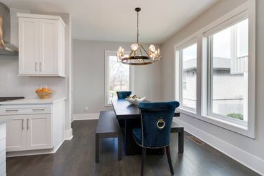 Open-concept modern dining area next to a white kitchen, dark hardwood floors, dark wood table with bench, blue velvet ring-back chairs for a pop of color, round brass chandelier, large bright windows.