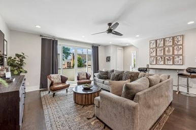 Bright modern living room with plush gray sectional, two leather accent chairs, round wooden coffee table on a patterned rug, ceiling fan, sliding glass doors to a balcony, botanical gallery wall and bar seating
