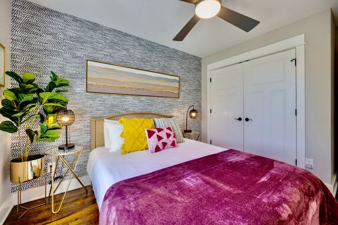 Cozy modern bedroom with a patterned accent wall, white bed topped with a magenta throw and yellow geometric pillows, potted fiddle-leaf fig in a gold planter, matching bedside lamps, ceiling fan, and white double closet.