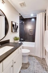 Bright modern bathroom with black elongated hexagon tile shower wall, white tub and curtain, white vanity with dark countertop, round mirror, patterned black-and-white floor tile and potted plant.