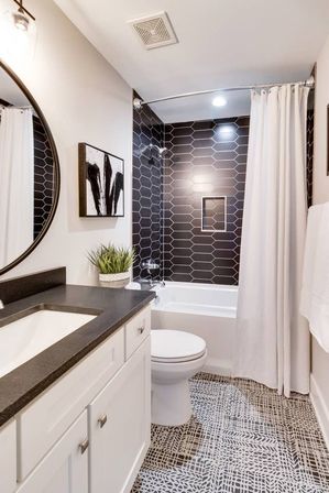 Bright modern bathroom with black elongated hexagon tile shower wall, white tub and curtain, white vanity with dark countertop, round mirror, patterned black-and-white floor tile and potted plant.