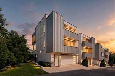 Contemporary three-story townhouses on a sloped street at sunset, vertical metal siding, glowing balconies and garage doors, concrete driveways and landscaped lawn.