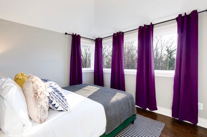 Bright modern bedroom interior with corner windows and deep purple curtains, white bedding, textured throw pillows, gray throw blanket and hardwood floor overlooking trees