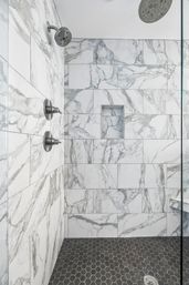 Sleek spa-like shower with white marble-look subway tiles and gray veining, recessed niche, stainless steel rain and wall showerheads, and dark hexagonal mosaic floor tiles.