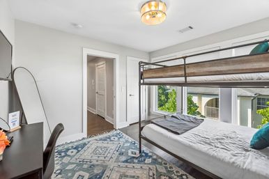 Bright, airy modern bedroom featuring a metal-frame bunk bed with wood accents, large windows with leafy outside view, patterned blue area rug, desk with mirror and hardwood floors.