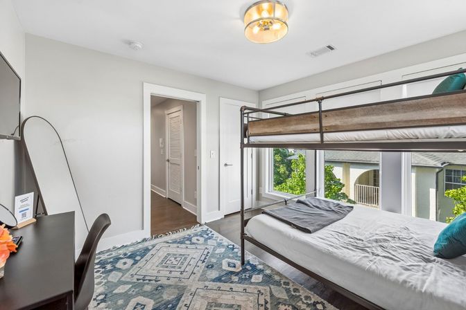 Bright, airy modern bedroom featuring a metal-frame bunk bed with wood accents, large windows with leafy outside view, patterned blue area rug, desk with mirror and hardwood floors.