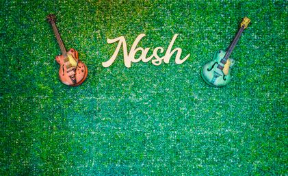 Music-themed green boxwood wall backdrop with white script sign reading "Nash" centered between a vintage orange guitar (left) and a teal guitar (right).