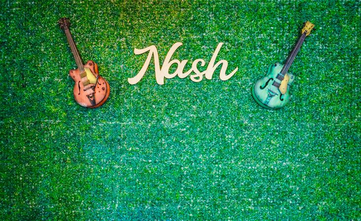Music-themed green boxwood wall backdrop with white script sign reading "Nash" centered between a vintage orange guitar (left) and a teal guitar (right).