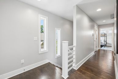 Sunlit modern upstairs hallway with dark hardwood floors, light gray walls, slim vertical windows and a white railing leading to a bedroom with a black metal bunk bed.