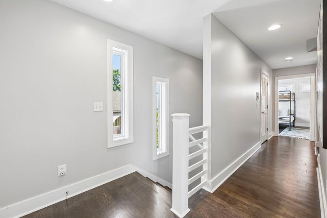 Sunlit modern upstairs hallway with dark hardwood floors, light gray walls, slim vertical windows and a white railing leading to a bedroom with a black metal bunk bed.