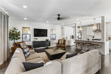 Bright open-concept living room and kitchen with a neutral gray sectional, leather accent chairs, dark wood coffee table, wall-mounted TV, white shaker cabinets and a long island with bar stools and glass pendant lights