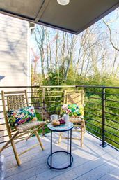 Cozy sunlit balcony nook with two woven wood chairs and colorful floral pillows, a small round bistro table holding coffee mugs, pastries and grapes, and a metal railing overlooking bare trees and green shrubs.