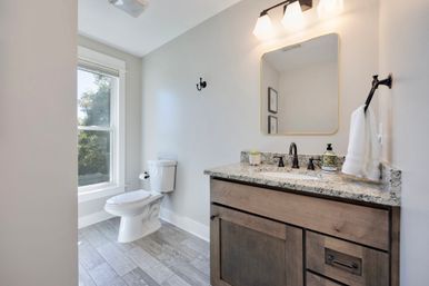 Sunlit modern residential bathroom with a white toilet by a large window, wood vanity with speckled granite countertop, black faucet and hardware, rectangular mirror, wall towel and gray wood-look tile floor.