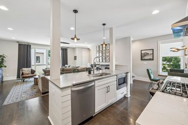 Modern open-concept kitchen and living room with white quartz island, stainless dishwasher and built-in microwave, pendant lights, dark hardwood floors, cozy sofa and armchair, and a sunlit dining nook by a window