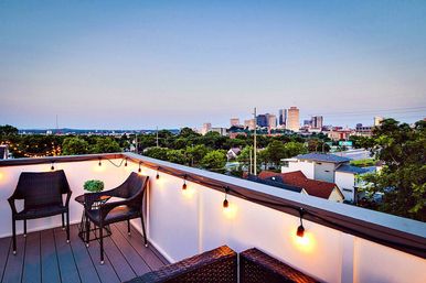 Cozy rooftop patio with wicker chairs, small bistro table, potted plant and warm string lights overlooking an urban city skyline at dusk