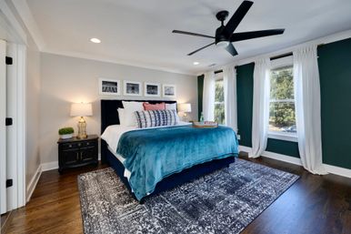 Spacious modern master bedroom interior with navy upholstered bed, teal velvet blanket, patterned area rug on dark hardwood floors, large sunlit windows with white curtains, black ceiling fan, and matching bedside lamps.