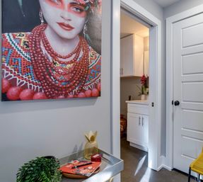 Contemporary home entryway with a large colorful portrait of a woman in red beads, gray walls, a tray with greenery and a gold bunny, and an open doorway to a small white-cabinet bathroom with a pink orchid.