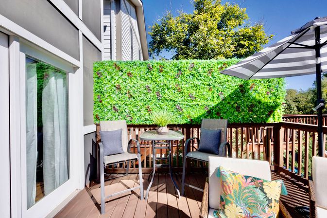 Sunny wooden backyard deck patio with sliding glass door, tall green privacy hedge, metal bistro table with potted plant, two cushioned bar chairs, striped patio umbrella and colorful throw pillow.