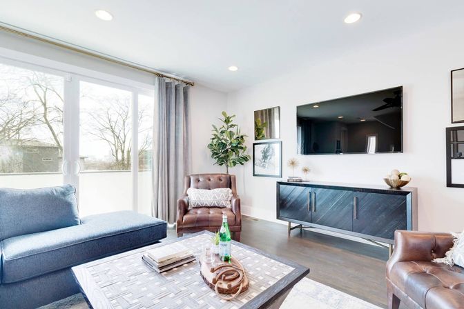 Sunlit modern living room with blue sectional, brown leather armchairs, patterned coffee table, wall-mounted TV, sliding glass doors, potted plant and hardwood floors.