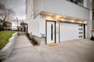 Contemporary white townhouse exterior with sleek glass-paneled entry door, attached garage with frosted windows, concrete driveway and side walkway bordered by a low retaining wall and shrubs under warm recessed lights.