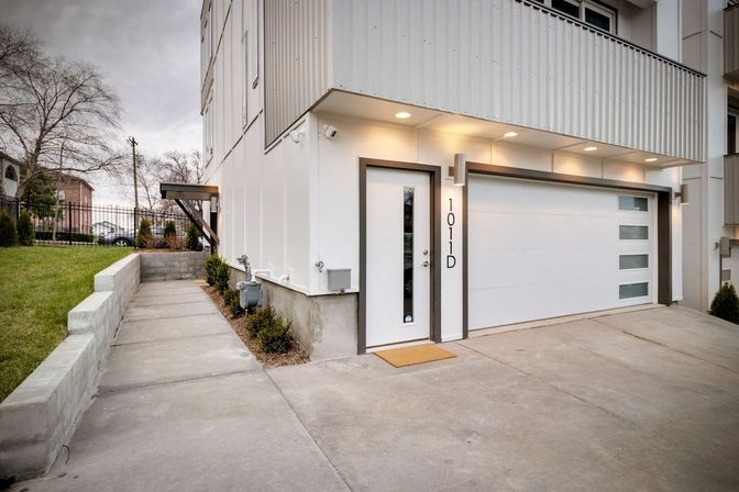 Contemporary white townhouse exterior with sleek glass-paneled entry door, attached garage with frosted windows, concrete driveway and side walkway bordered by a low retaining wall and shrubs under warm recessed lights.