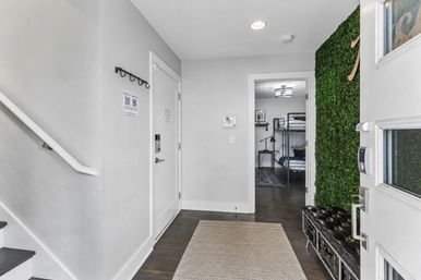 Modern home entryway with dark hardwood floors and patterned runner, white staircase on left, tufted leather bench and green living-wall panel by the door, keypad lock and wall security panel, open doorway showing a bedroom with metal bunk beds.