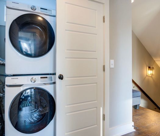 Stacked front-loading washer and dryer tucked behind a white paneled closet door in a modern home hallway with hardwood floors and stairwell light