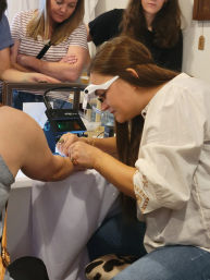 Jewelry artisan wearing a magnifying visor inspects and adjusts a bracelet on a customer's wrist at a craft market table while shoppers watch.