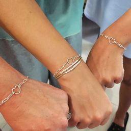 Close-up of three wrists wearing matching silver heart-link chain bracelets, center wrist with stacked gold and pearl bracelets — cute fashion jewelry close-up.