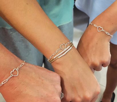 Close-up of three wrists wearing matching silver heart-link chain bracelets, center wrist with stacked gold and pearl bracelets — cute fashion jewelry close-up.