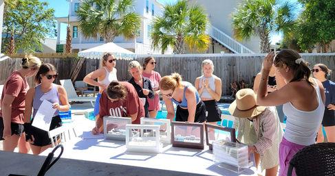 Poolside scene of a group of women browsing jewelry display cases and signing forms at a sunny beach-house backyard with palm trees