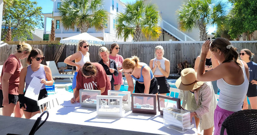 Poolside scene of a group of women browsing jewelry display cases and signing forms at a sunny beach-house backyard with palm trees