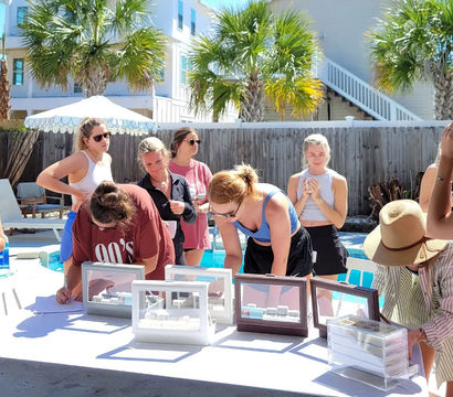 Poolside scene of a group of women browsing jewelry display cases and signing forms at a sunny beach-house backyard with palm trees